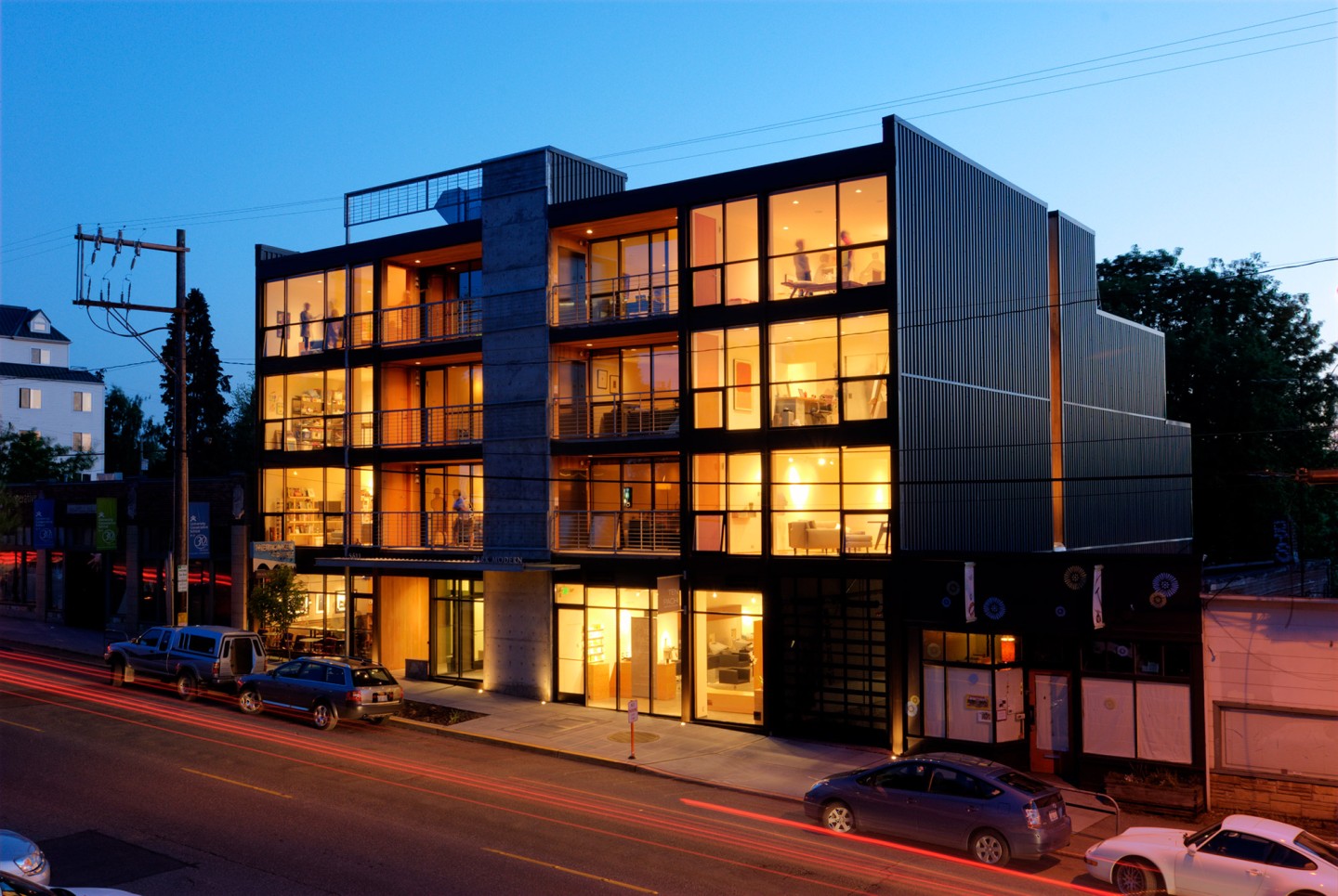  Park Modern. Photograph courtesy of BUILD LLC. Four-story building at dusk on the side of a city street. Large windows with lit units are visible, with brightly lit rooms and people in some of the units.