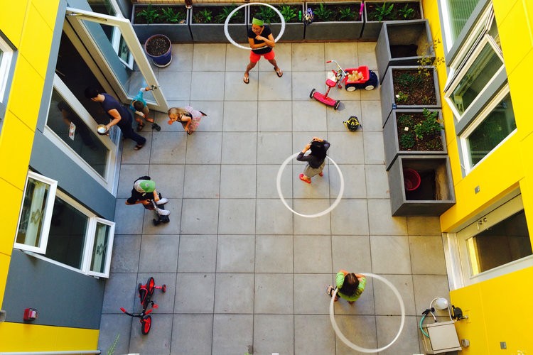 CHUC, courtyard. Photographs courtesy of Schemata Workshop/ William Wright Photography. Overhead photo of people gathered on a open-air commons area, hoola-hoopin'. Some planters are on the side, and some kids toys are scattered in the corner nearby,